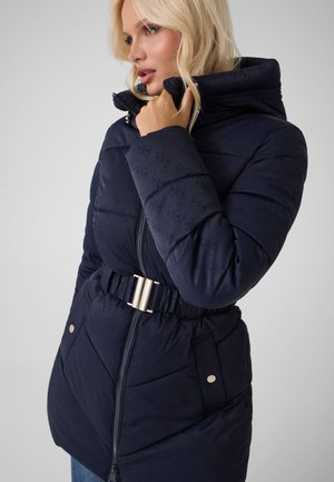 Mujer rubia con chaqueta acolchada azul marino con cuello alto y cinturón con hebilla plateada, mirando hacia un lado.