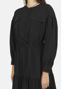 Robe noire à manches longues avec un col haut, à boutons devant, deux poches poitrine, une taille élastique et un bas en couches. Tissu lisse et manches froncées.