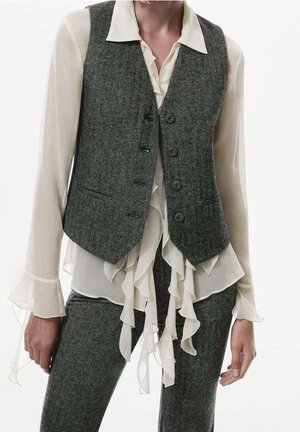 Femme portant un gilet en laine gris et un pantalon assorti sur un chemisier blanc transparent à volants avec col pointu et poignets évasés.