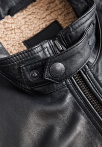 Gros plan du col d'une veste en cuir noire avec doublure en polaire beige, bouton-pression métallique marqué « Signature Durable Goods » et fermeture éclair visible.