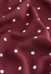 Tissu bordeaux avec une texture lisse et de petits pois blancs régulièrement espacés, présenté avec de doux plis et un éclairage doux.