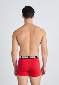Boxeri scurți bărbați roșii din material elastic, cu un brâu negru în care este inscripționat cu alb „PUMA”. Vederea din spate arată un design ajustat și picioare scurte.