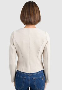 Giacca beige strutturata con maniche lunghe e scollatura rotonda, caratterizzata da dettagli delle cuciture e una silhouette aderente, indossata sopra jeans blu.