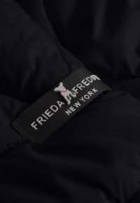 Zwart donzig stof met een gelabelde zwarte label met "FRIEDA & FREDDY" en een zilveren honden silhouet met een rode halsband.