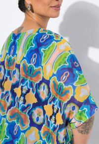 Blusa colorata con motivo floreale nelle tonalità di blu, verde, giallo e rosa. Tessuto leggero con una vestibilità ampia e maniche corte.