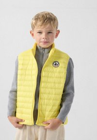 Gele puffer-vest met een doorgestikt ontwerp, ritssluiting en een rond logo, gedragen over een grijze sweater met rits. Gestructureerde stof en moderne snit.