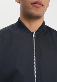 Chaqueta bomber negra con cuello acanalado, cierre de cremallera completa y tejido suave. Cuenta con un tirador de cremallera plateado con detalle de logo.