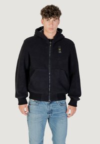 Felpa nera con zip, dotata di tasche frontali e polsini a coste. Il tessuto ha una consistenza morbida. Indossata con jeans di denim blu chiaro.