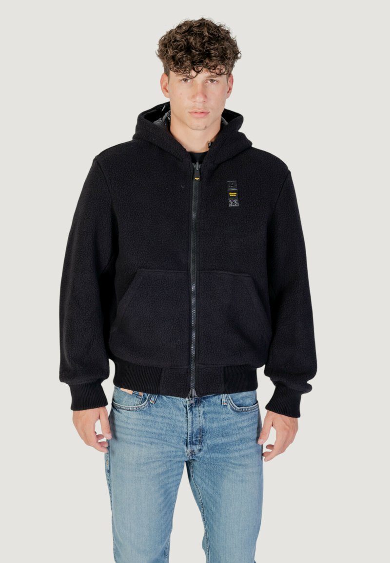 Felpa nera con zip, dotata di tasche frontali e polsini a coste. Il tessuto ha una consistenza morbida. Indossata con jeans di denim blu chiaro.