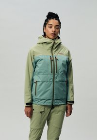 Giacca impermeabile di colore verde chiaro con un pannello frontale verde scuro. Presenta diverse tasche con zip e un cappuccio regolabile.