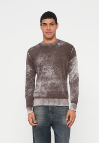 Mann trägt einen braun-weiß marmorierten Pullover mit langen Ärmeln und ausgewaschene blaue Jeans, steht vor einem schlichten weißen Hintergrund.