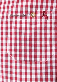 Rood-wit geruit stof met een knoop en een geborduurd rendierlogo boven de borstzak; gladde textuur, nauwsluitende pasvorm.