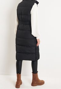 Doudoune sans manches noire avec une taille ceinturée, col montant et coutures verticales, portée avec des leggings noirs et des bottines marron.