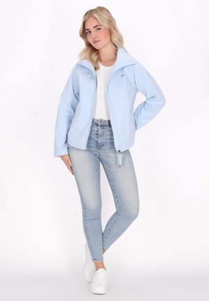 Blonde vrouw met een lichtblauw jasje met rits, wit shirt, lichtblauwe skinny jeans en witte sneakers, staand met één hand op de heup.