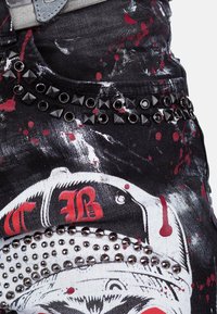 Pantaloni di denim neri con schizzi rossi, con borchie argento e nere sulla vita. Stampa grafica notevole sul tessuto.