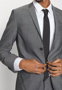 Szara, dopasowana marynarka na jeden czarny guzik, w połączeniu z białą koszulą i czarnym krawatem, prezentująca gładką fakturę.
