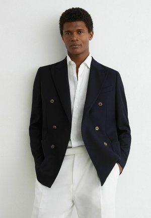 Man wearing a black double-breasted blazer over a white buttoned shirt and white pants standing against a white wall.