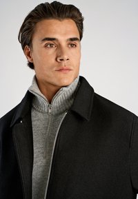 Svart ullrock med slagkrage över en grå stickad polotröja. Detaljer inkluderar en silverfärgad blixtlås framtill.