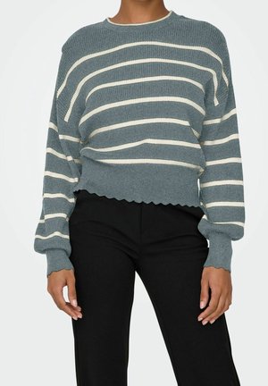Pull tricoté rayé bleu et blanc avec ourlet et poignets festonnés porté avec un pantalon noir sur une personne debout.