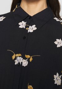 Schwarzes Hemd mit Knöpfen und floralem Muster in Weiß und Gelb. Glattes Material mit spitzem Kragen und sichtbaren Knöpfen.