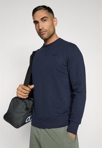 Felpa blu navy in misto cotone con colletto a girocollo, polsini e orlo a costine. Dettaglio logo nero sul petto. Il modello tiene in mano una piccola borsa nera.