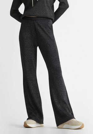 Osoba ubrana w ciemnoszare, teksturowane spodnie o szerokich nogawkach oraz dopasowaną bluzę z kapturem, do tego beżowo-białe trampki na jasnym tle.