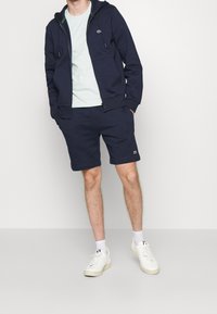 Sudadera con cremallera en color azul marino con un acento verde bajo la capucha, combinada con pantalones cortos a juego en azul marino. Zapatillas blancas completan el atuendo.