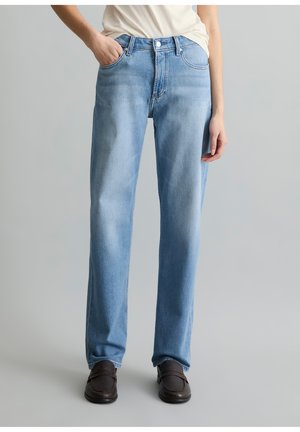 Person, der hellblaue Jeans mit geradem Bein, ein weißes Hemd und dunkelbraune Loafers vor einem schlichten grauen Hintergrund trägt.