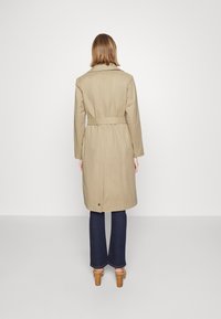 Béžový trench coat s opaskem v pase, dlouhými rukávy a detailem ventilu na zadní straně. Kombinovaný s modrými zvonovými džíny a béžovými sandály na podpatku.