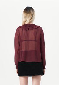 Burgunderfarget gjennomsiktig bluse med volangkrage, lange ermer og smockdetaljer ved halsen. Kombinert med et svart skjørt som har lagdelt design.