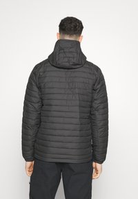 Svart puffjacka med quiltat mönster, fast huva och elastiska ärmmuddar. Lättvikts, mjukt texturerat tyg. Bakvy som visar axelsömmarna.