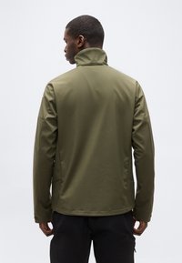 Homme portant une veste col montant vert olive, vu de dos, debout devant un fond clair uni.
