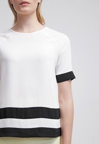 Haut blanc à manches courtes avec des rayures noires horizontales à l'ourlet et aux manches. Texture lisse avec une coupe décontractée ; encolure ronde.