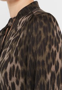 Blusa a maniche lunghe in tessuto leggero e trasparente con stampa leopardata marrone e nera. Presenta un colletto alto e un dettaglio con fiocco al collo.
