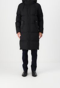 En person klädd i en lång svart vadderad kappa med huva, mörka byxor och svarta finsskor, står mot en enfärgad vit bakgrund.