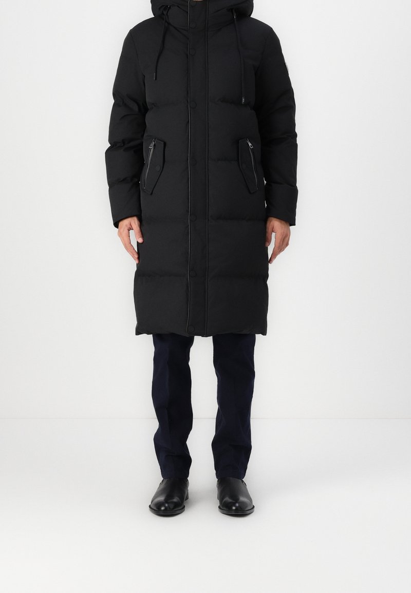 En person klädd i en lång svart vadderad kappa med huva, mörka byxor och svarta finsskor, står mot en enfärgad vit bakgrund.