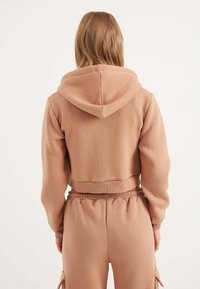 Hoodie accorciato in morbido tessuto marrone chiaro, con cappuccio, caratterizzato da polsini e orlo a coste. Abbinato a pantaloni rilassati dello stesso tessuto.
