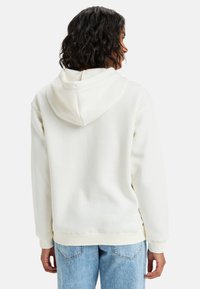 Sudadera blanca con capucha, hecha de un tejido texturizado. Cuenta con una capucha ajustable y puños acanalados. La vista trasera muestra un corte relajado. Combinada con jeans de color azul claro.