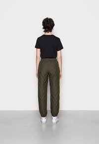 Zwarte short-sleeve shirt, olijfgroene gewatteerde broek met gele zijstreep, en witte sneakers. Effen achtergrond. Achteraanzicht.