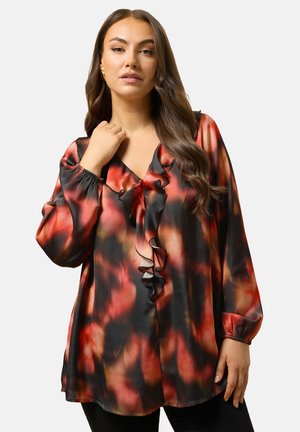 Nainen, jolla on pitkät ruskeat hiukset, pukeutuneena löysään punamustaan kuviolliseen paitaan, jossa on röyhelöinen V-pääntie ja pitkät hihat. Hän poseeraa yksi käsi kaulusta lähellä.