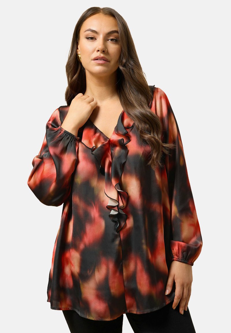 Kvinde med langt brunt hår iført en løs rød og sort mønstret bluse med v-udskæring og lange ærmer, poserende med den ene hånd nær kraven.