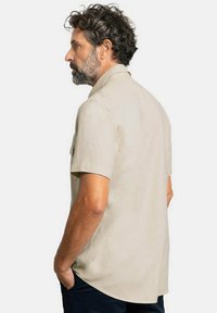 Camisa beige de manga corta con una textura suave, dos bolsillos en el pecho y un dobladillo recto, llevada por un hombre de espaldas a la cámara.