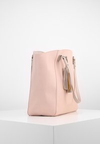 Sac fourre-tout en cuir rose clair avec une bandoulière unique et des glands décoratifs, exposé sur un socle blanc sur fond uni.