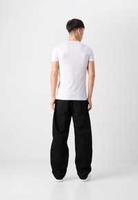 T-shirt bianco aderente, maniche corte, texture liscia, abbinato a pantaloni neri larghi con tasche posteriori, indossato con sneakers bianche.