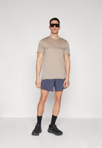 Beige T-shirt met korte mouwen en een subtiele horizontale grafische print, gecombineerd met marineblauwe shorts. Het model draagt zwarte zonnebril en sneakers.