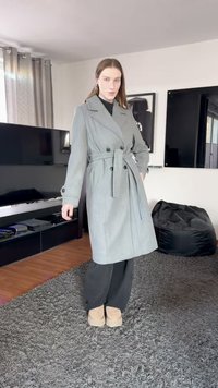 Pelēks dubultpogājams trančmētelis ar jostu, ar lielām pogām un izstrādātu dizainu. Valkāts virs melnas apģērba un bēšām zābakām.