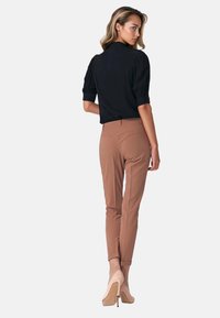 Kobieta z długimi włosami, ubrana w czarną bluzkę z bufkami, brązowe dopasowane spodnie i beżowe wysokie obcasy, stoi tyłem.