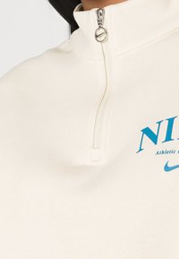 Krämfärgad zip-up sweatshirt med hög krage, utrustad med en turkos Nike-logga och text. Tillverkad i mjukt tyg med en slät struktur.