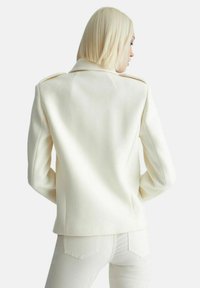 Giacca bianca su misura con spalle strutturate e una texture liscia. La vista posteriore rivela linee pulite e dettagli di design minimi.