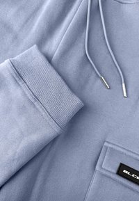 Close-up van de lichtblauwe sweatshirtmouw, trekkoorden met metalen uiteinden en een zak met een zwart-wit rechthoekig label.